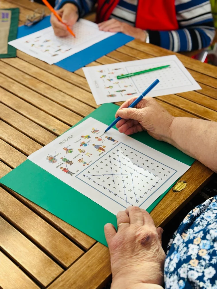 Senioři u stolu vyplňují pastelkami pracovní listy s osmisměrkou